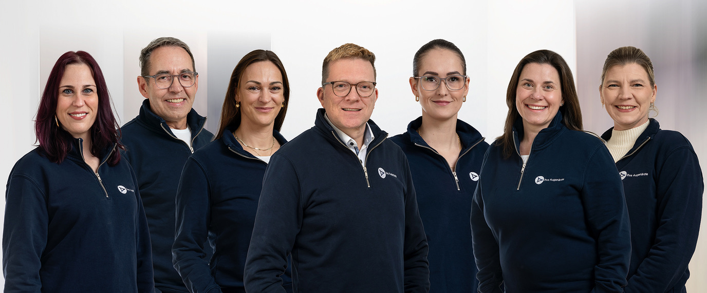 Sieben Personen in dunkelblauen Pullovern lächeln freundlich in die Kamera vor einem hellen Hintergrund.