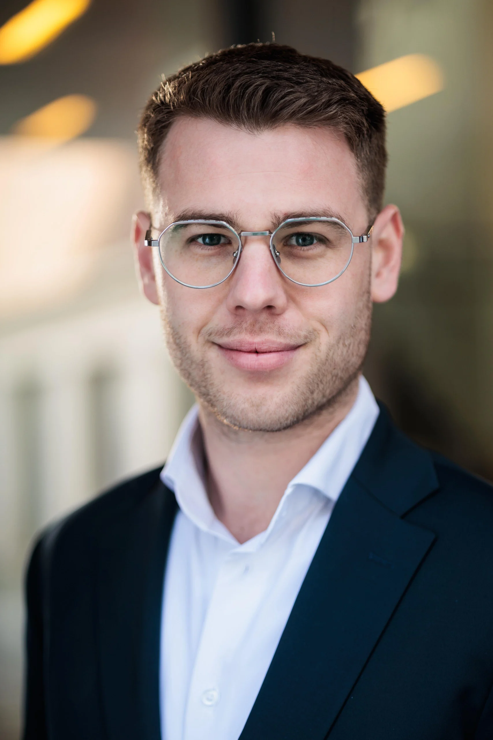 Porträt eines lächelnden Mannes mit Brille im Anzug vor unscharfem, neutralem Hintergrund.