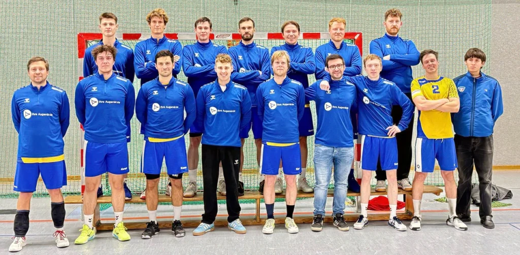 Eine Gruppe von Handballspielern in blauen Trikots posiert lächelnd vor einem Hallentor.