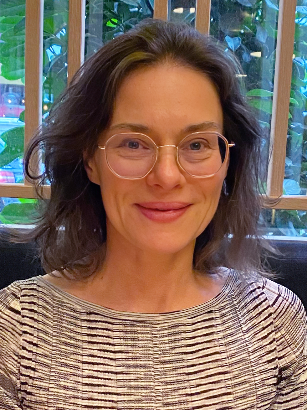 Eine lächelnde Person mit Brille vor grünem Hintergrund, trägt ein gestreiftes Oberteil.