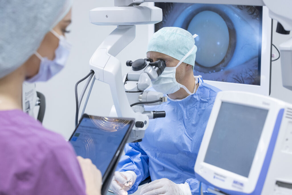 Chirurg operiert Auge mit Mikroskop, Assistentin dokumentiert auf Tablet, Bildschirm zeigt vergrößertes Auge.